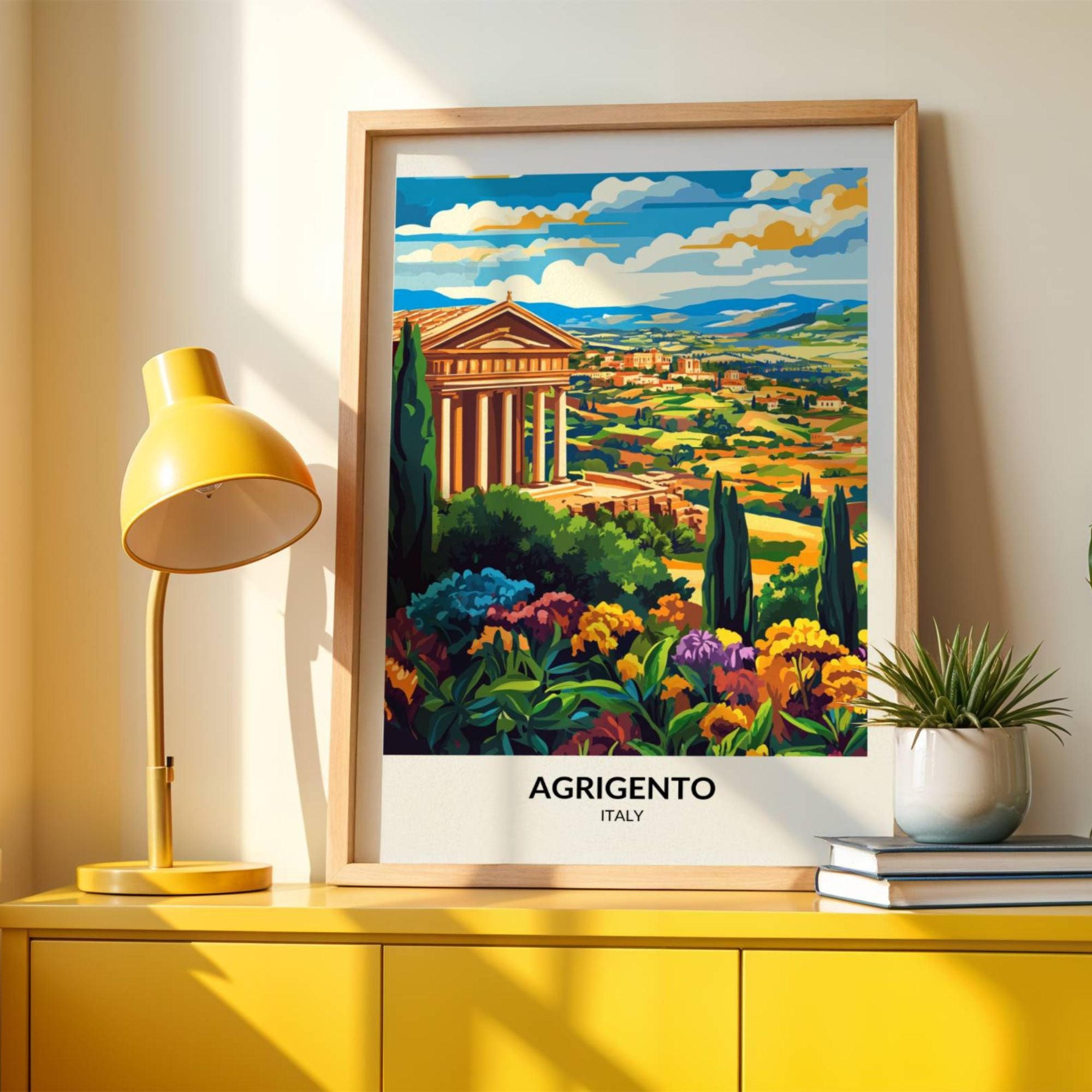 Framed travel poster of Agrigento, Italy, featuring a vibrant illustration of the Valley of the Temples with ancient Greek ruins, lush colorful flowers, and rolling countryside in the background. The poster is displayed on a modern yellow cabinet, accompanied by a matching yellow lamp and a small potted plant, with soft natural light casting shadows on the wall.