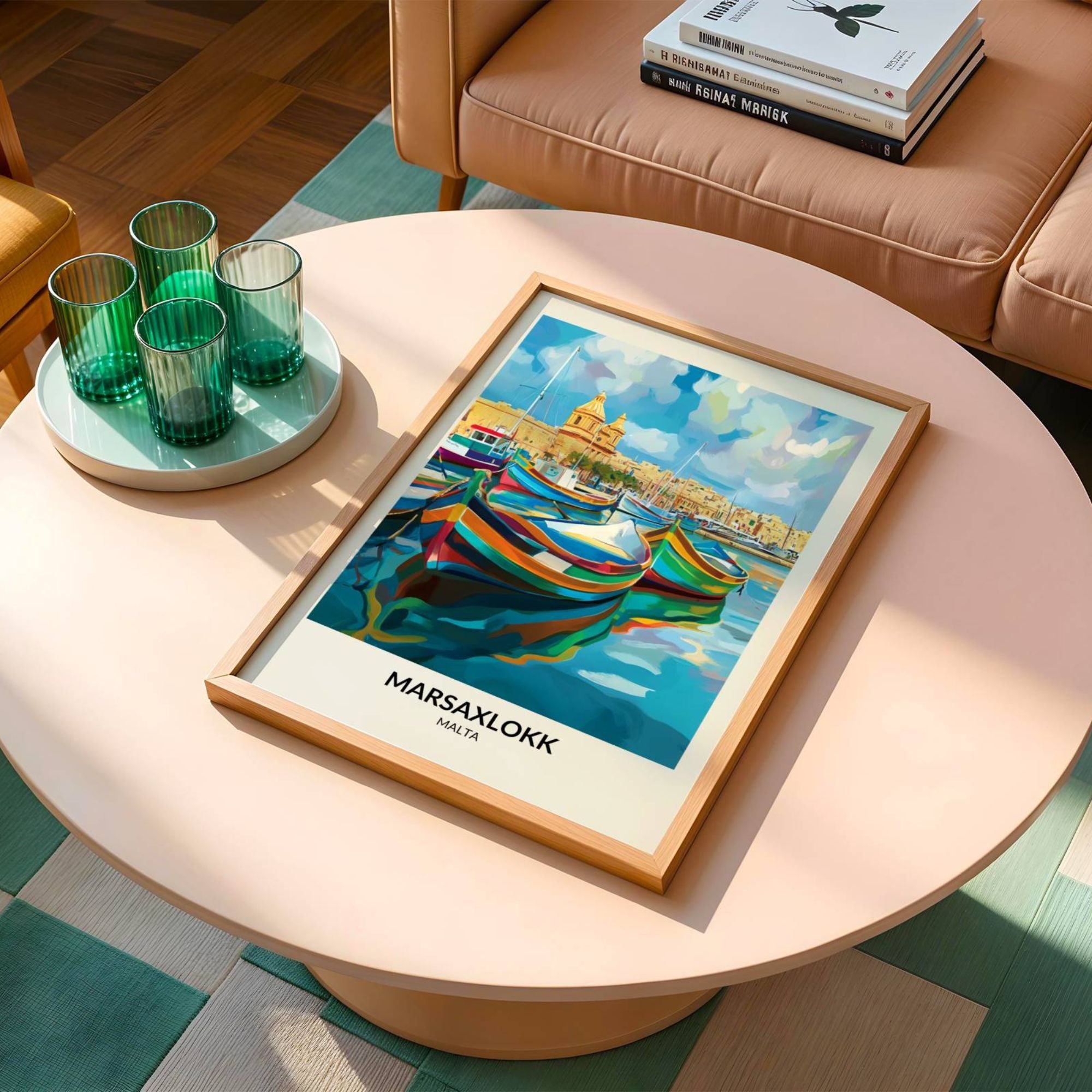 Framed travel poster of Marsaxlokk, Malta, featuring a vibrant illustration of traditional Maltese fishing boats floating on calm blue waters with a historic townscape in the background. The poster is placed on a stylish pastel pink coffee table, accompanied by green glassware and a modern beige sofa with books stacked on the armrest.